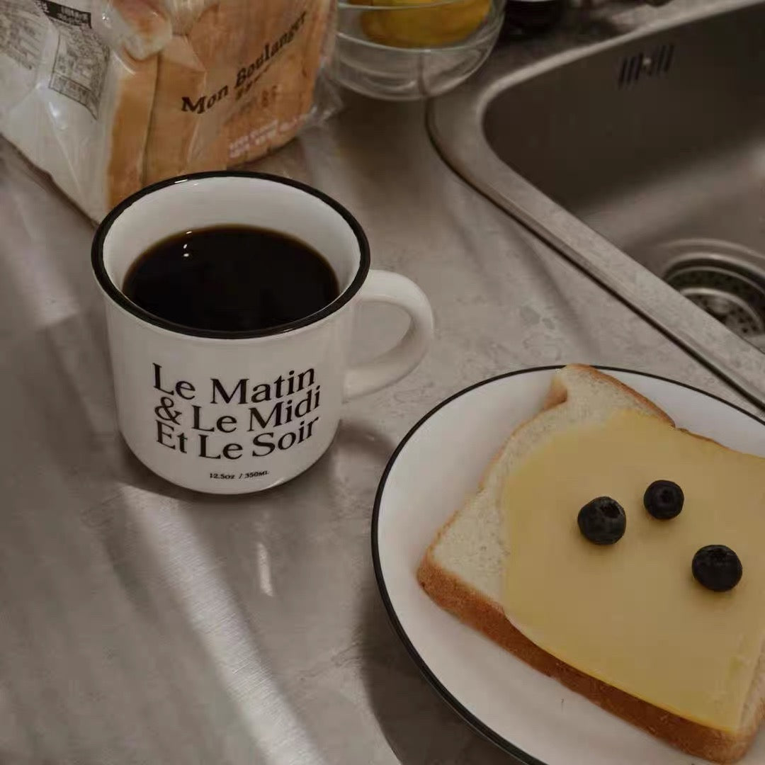 A white enamel coffee mug with black text reading 'Le Matin & Le Midi Et Le Soir' sits on a kitchen counter next to a plate of cheese and blueberry toast, with a bag of bread and a sink in the background.