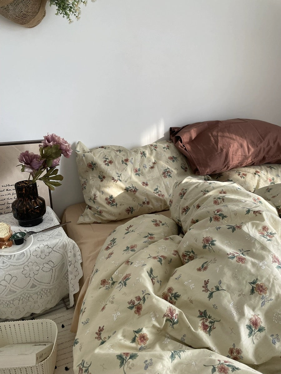 Close view of vintage floral bedding beside rust pillows, flowers, framed art, and a lace-covered table.
