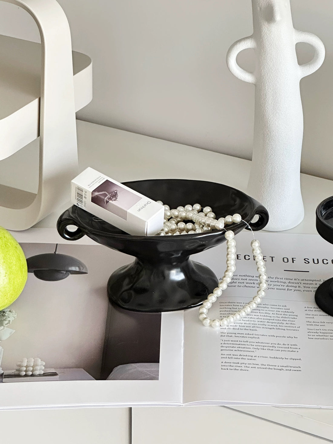 A silver metallic bowl-shaped tray holding an open invitation card and a wristwatch, placed on an open book on a wooden table. A pair of silver over-ear headphones sits in the background.