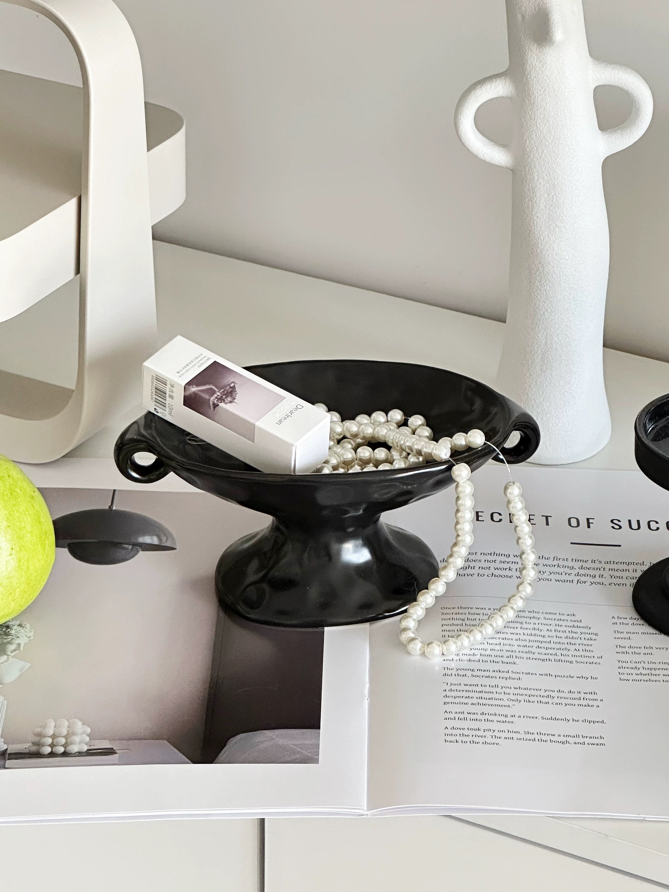 A black pedestal bowl with side handles, styled on a white surface with a white pearl necklace, a small white box, and an open magazine underneath. A white sculptural vase stands behind it, and part of a green fruit is visible on the left.