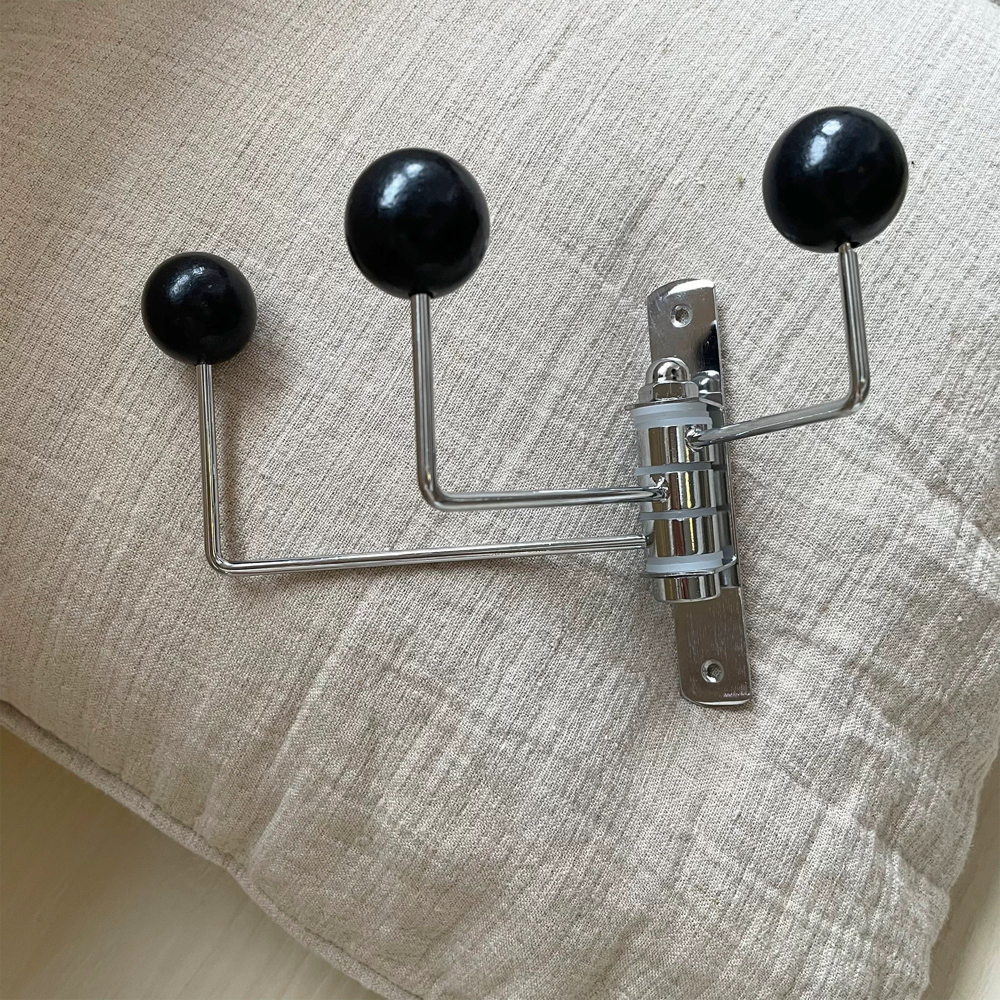 A three-hook wall coat rack with black spherical handles and a chrome metal mounting bracket, shown resting on a textured beige fabric surface.