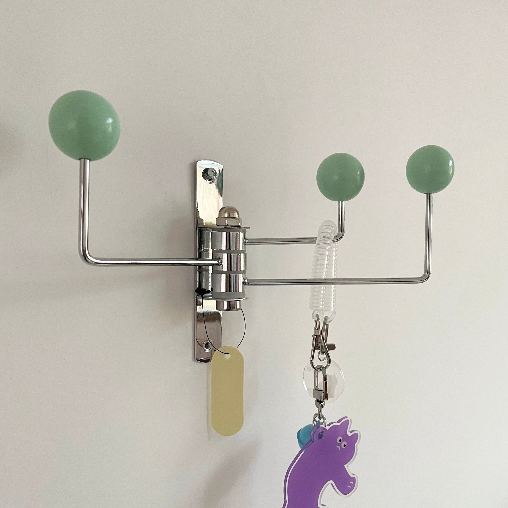 A wall-mounted metal key rack with three spherical green hooks and hanging keys and tags against a plain light wall.