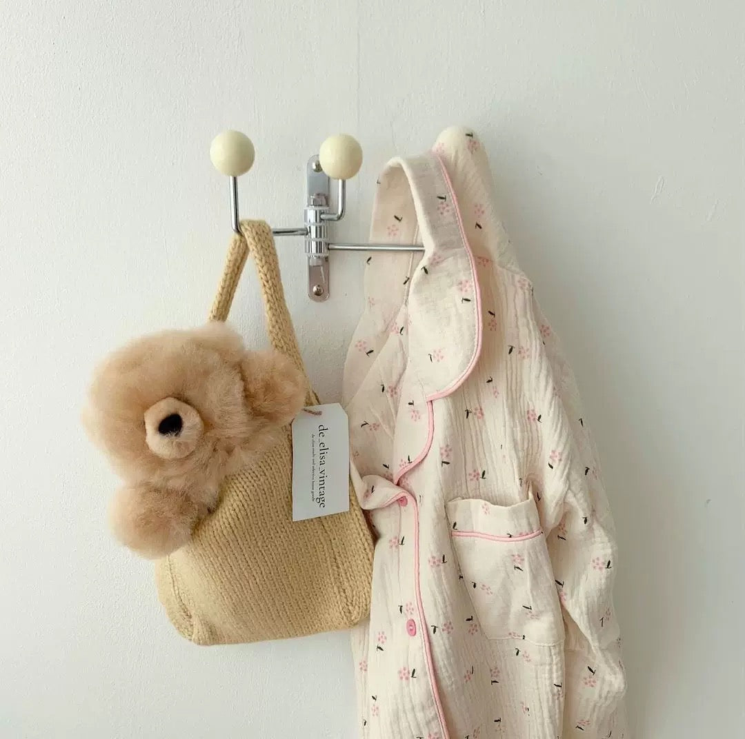A cream-colored cotton muslin pajama set with pink trim hangs on a wall-mounted metal hook next to a beige woven tote bag and a fluffy beige teddy bear.