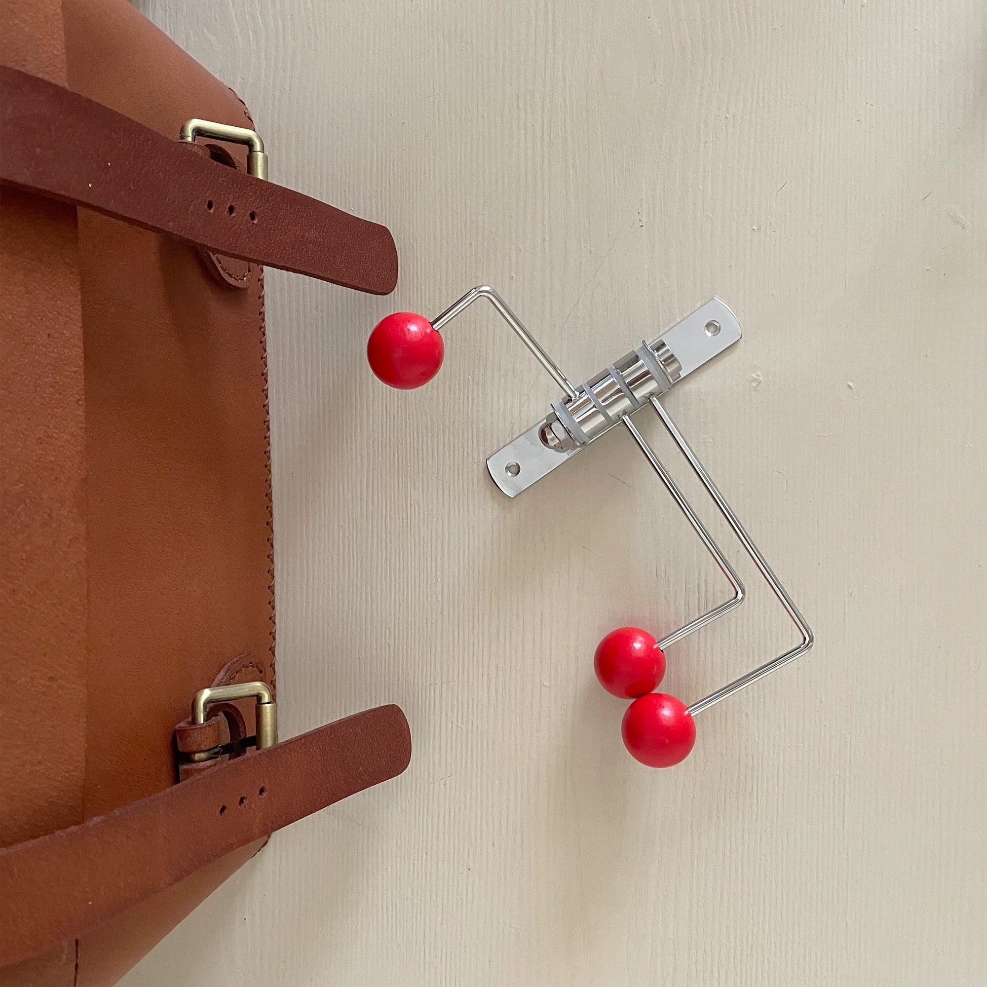 A close-up of a brown leather bag strap next to a silver metal wall hook with three red spherical beads, mounted on a light beige textured wall.
