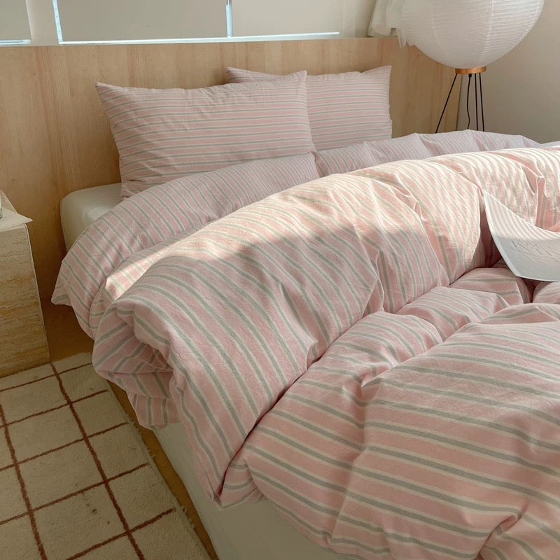 Bed with pink and white striped bedding in a room with wooden flooring and a lamp.