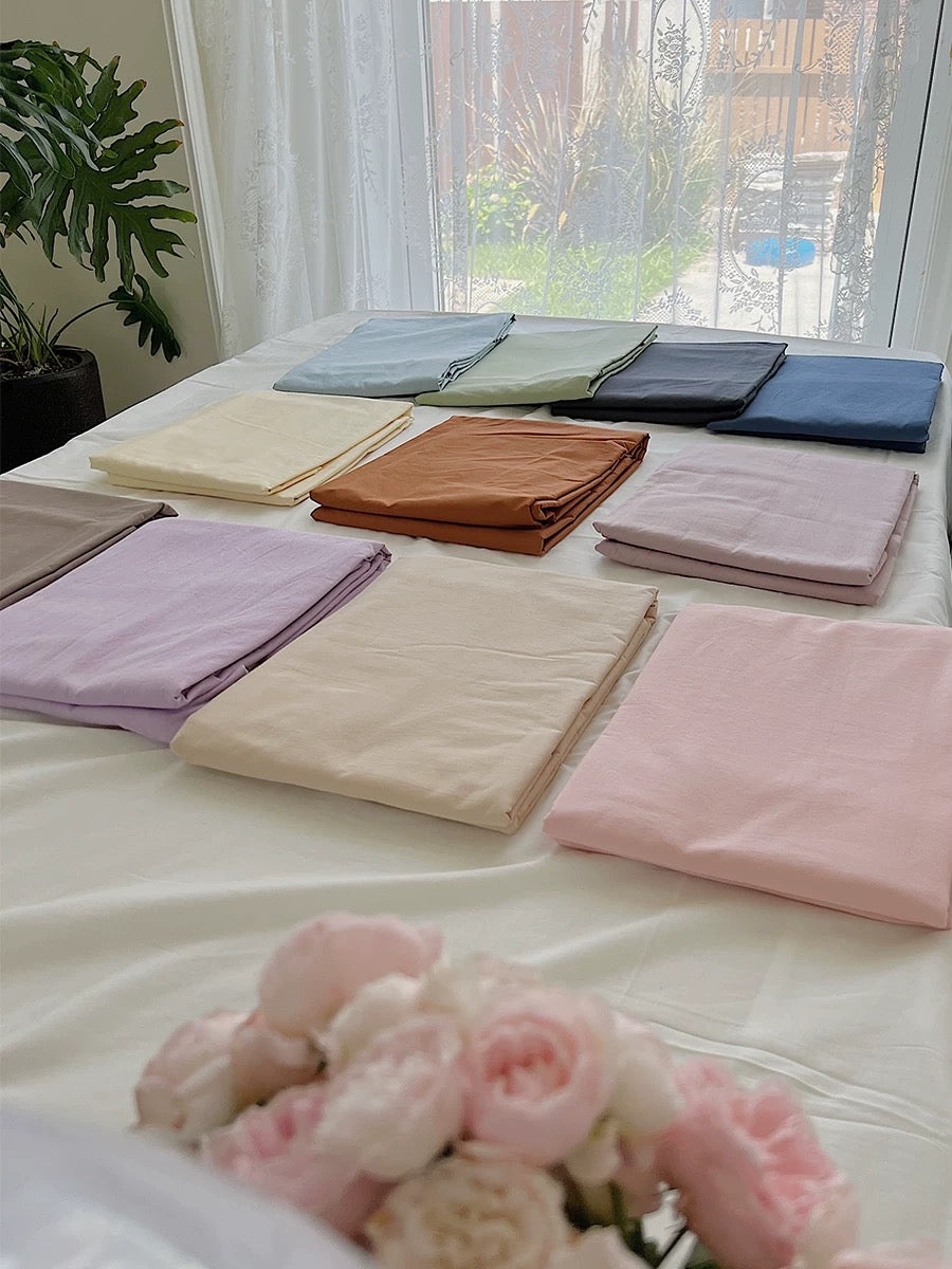 Folded solid-color sheets are spread across a bed beside a bright window and pink flowers.