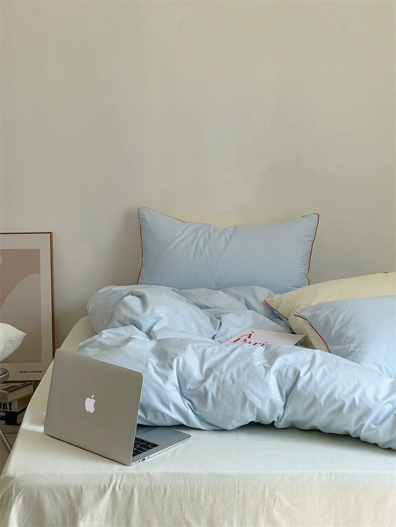 Laptop on a bed with light blue bedding and pillows against a plain wall.