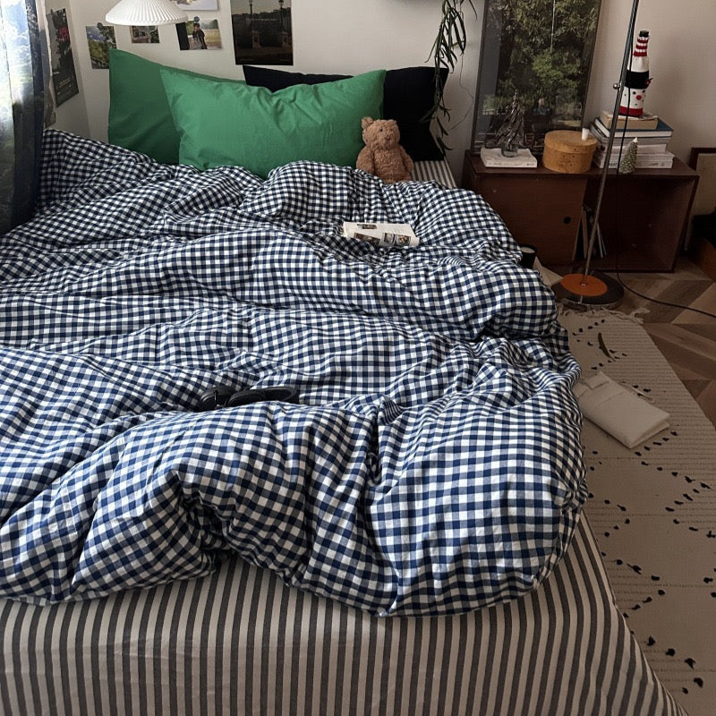 A bed in a real bedroom setting with a blue and white gingham checkered duvet cover and matching striped bedding, styled with green and black pillows, a teddy bear, and bedside furniture.
