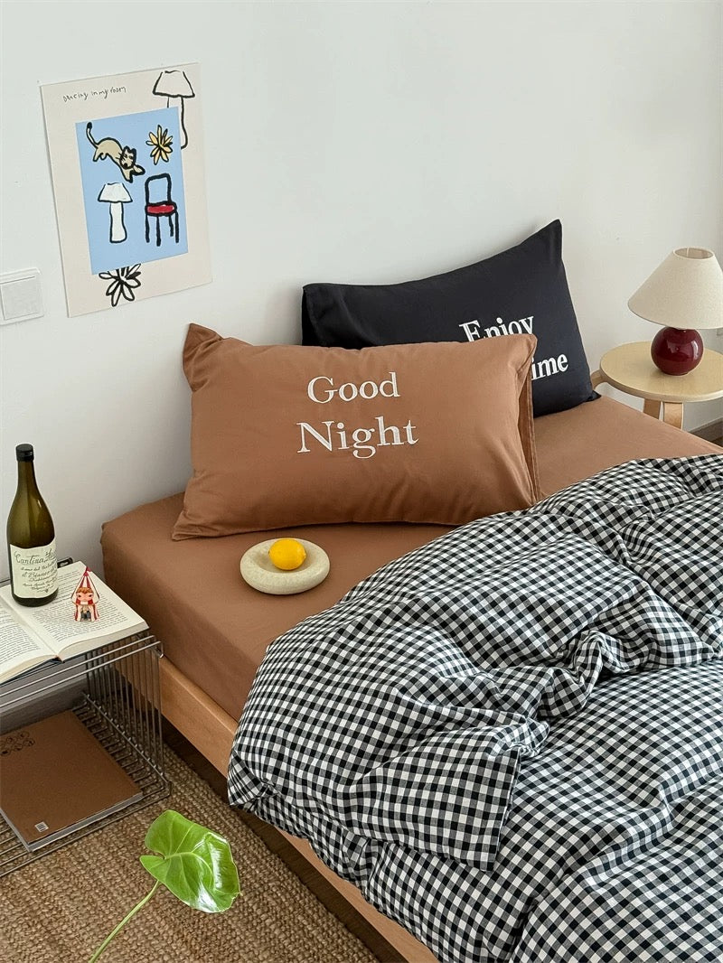 Bedroom with a bed featuring a 'Good Night' pillow and checkered blanket, with a bottle and small plant on the floor.