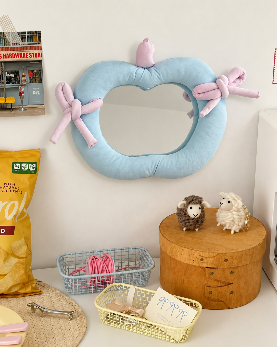 A heart-shaped light blue cushioned mirror with pink bow details on the sides, displayed on a white wall in a styled room setting with a wooden box, small sheep figurines, and baskets.