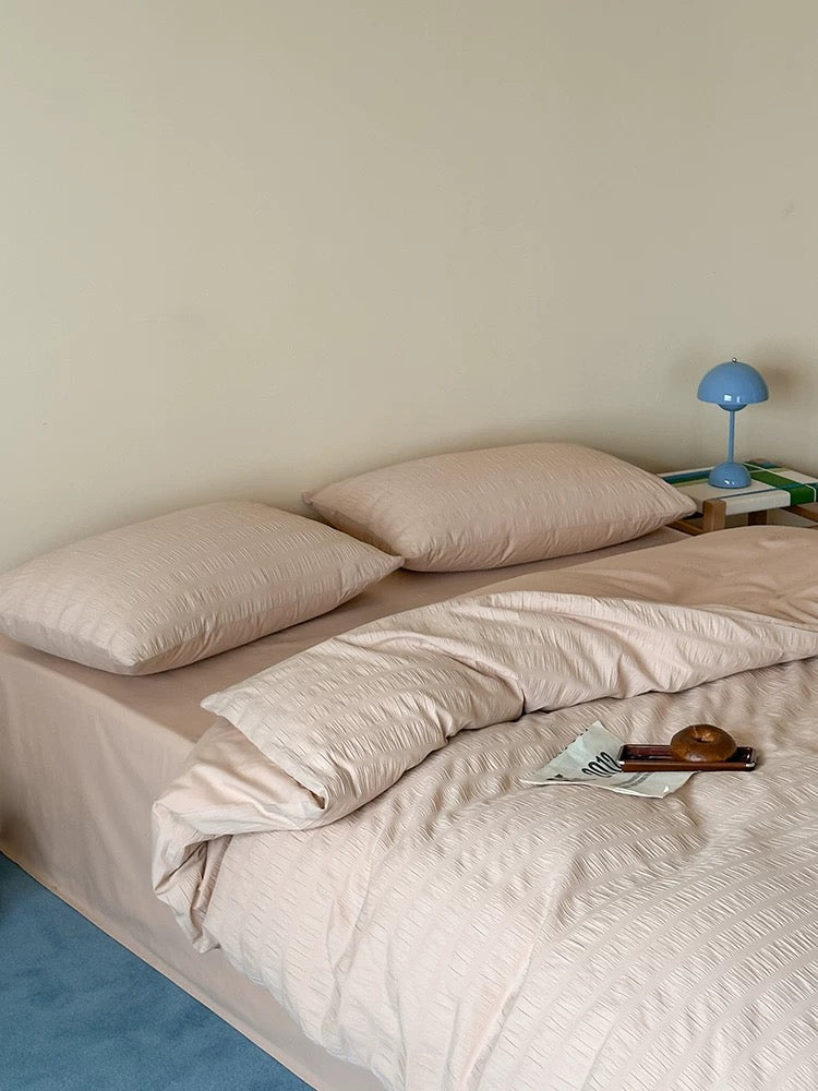 A neatly made bed with blush pink textured bedding, including two pillows, a matching duvet cover, and a sheet, set against a plain beige wall with a small blue bedside lamp.