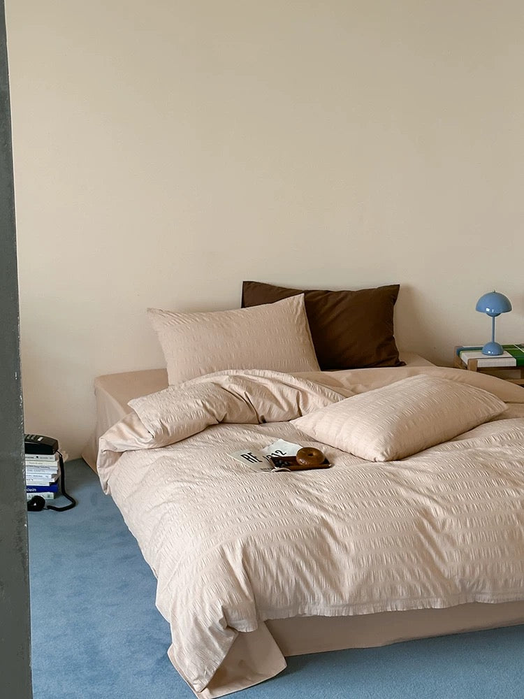 A neatly made bed with light beige textured bedding, including a duvet cover and two matching pillows, styled in a minimalist bedroom with a blue carpet, cream wall, and a small bedside lamp.