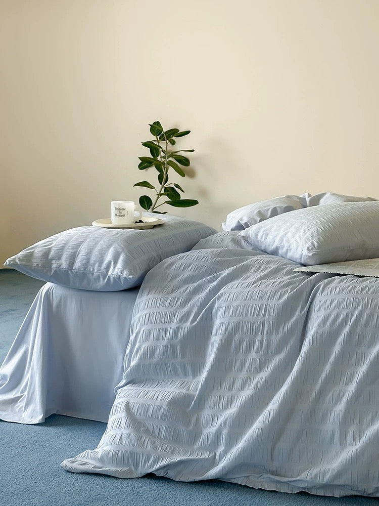 Light blue crinkled bedding set including a duvet cover and matching pillowcases, styled on a bed in a minimalist bedroom with a beige wall and blue carpet.