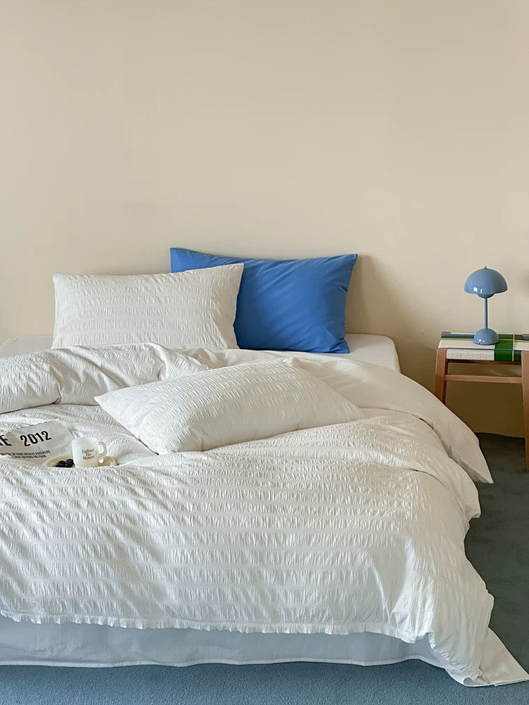 A neatly made bed with white textured bedding and two pillows, one white and one blue, in a minimalist bedroom setting with a beige wall and a small wooden nightstand.