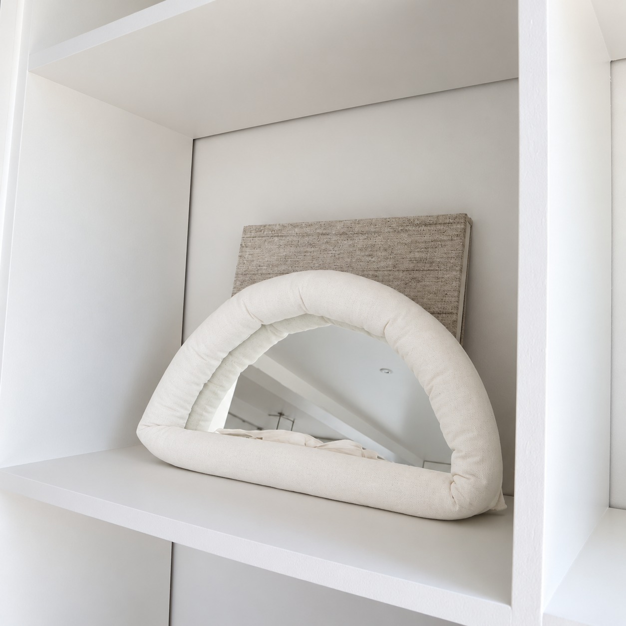 A cream-colored, arched cushioned mirror with a thick padded frame, displayed on a white shelf next to a textured beige canvas print.