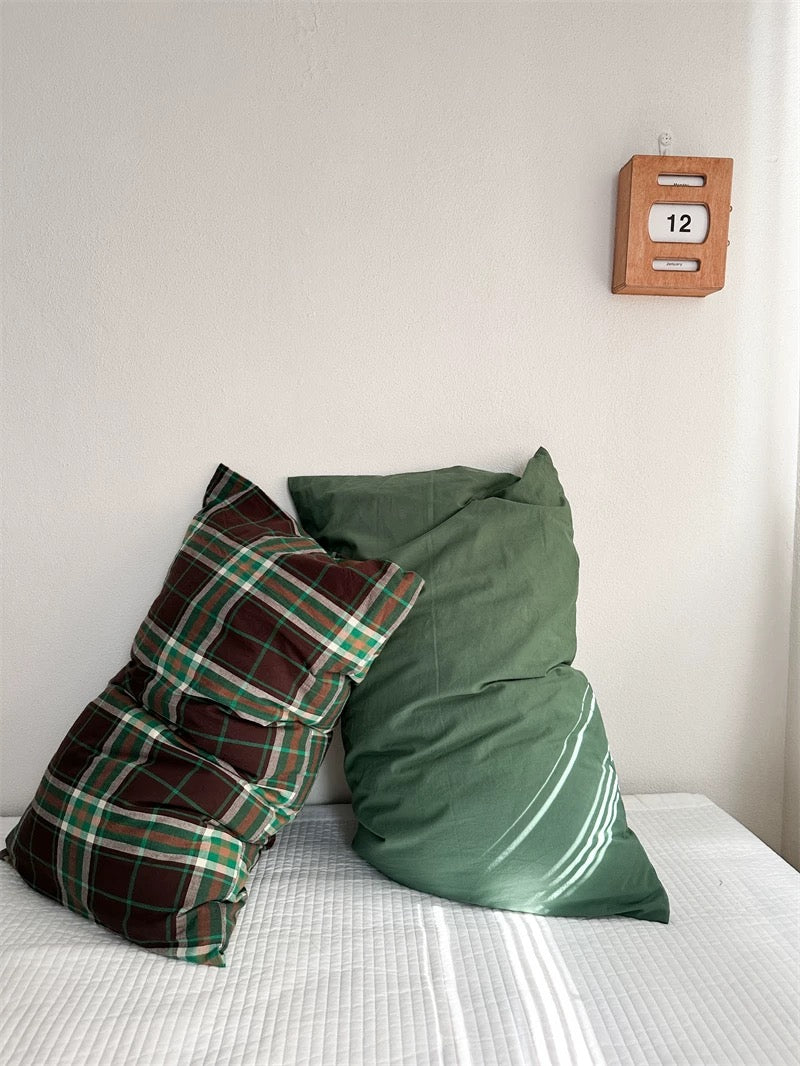 A green pillowcase sits beside a brown and green plaid pillowcase, with another green pillowcase behind them.