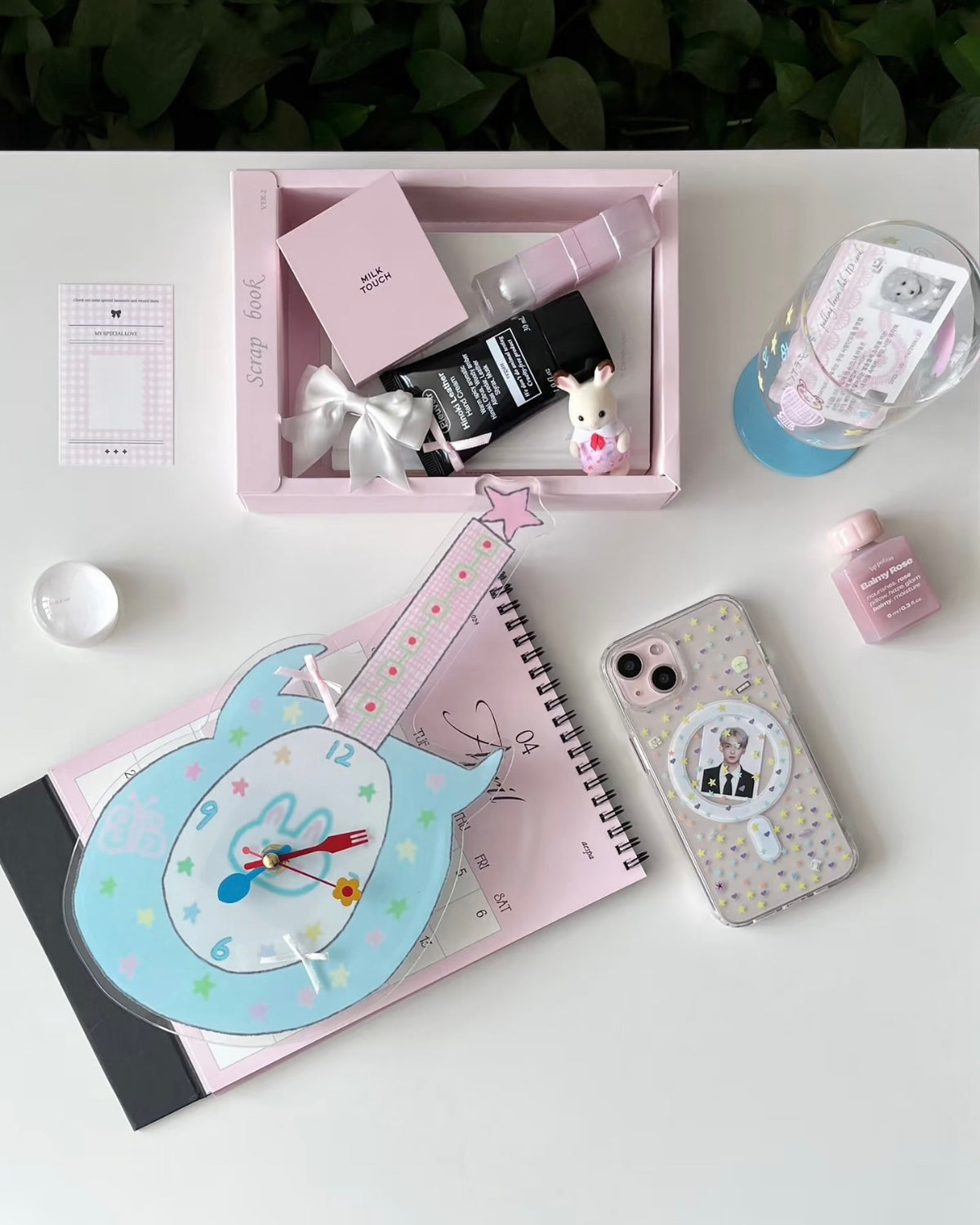A styled flat-lay of pastel pink stationery and beauty accessories on a white surface, including a pink tray with cosmetics, a guitar-shaped clock, a glittery phone case, a notebook, and small decorative items.