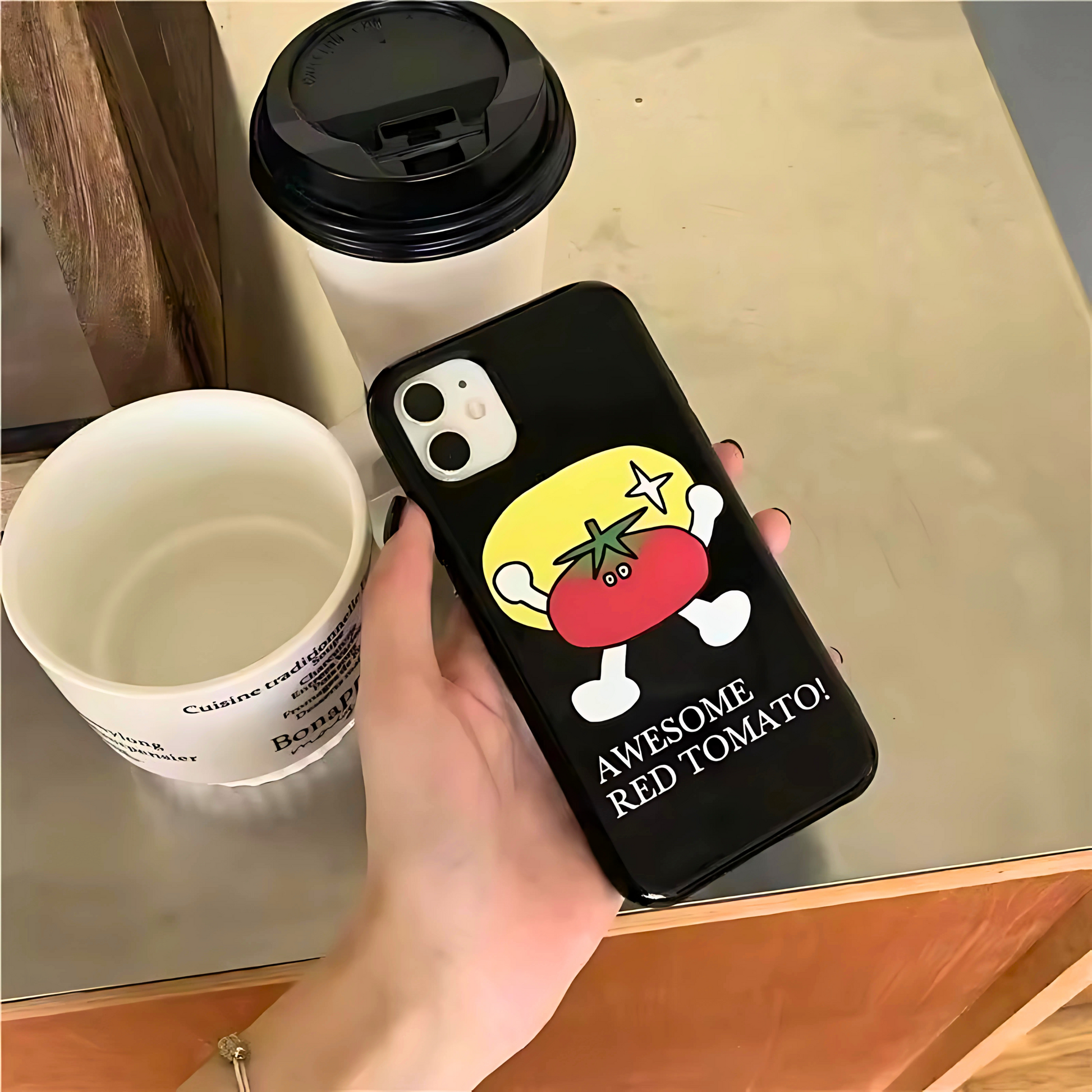 Phone case with cartoon tomato design and text 'Awesome Red Tomato' held by a hand next to a coffee cup.