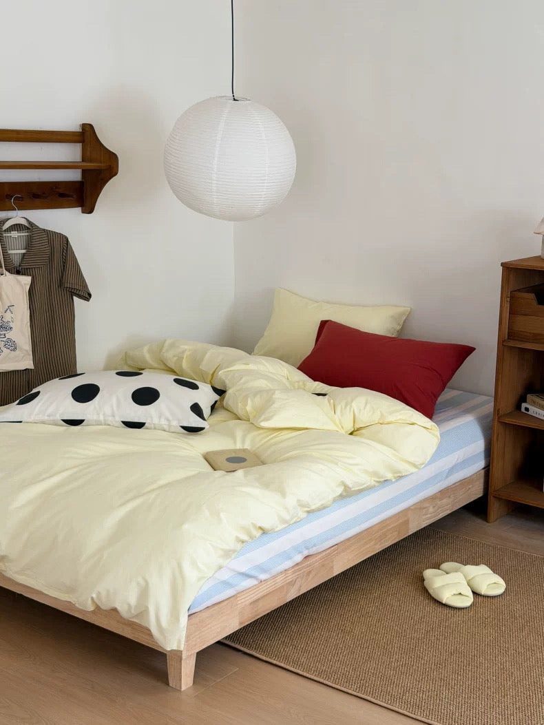 A cozy bedroom scene with a light wood bed frame dressed in pale yellow bedding, a red accent pillow, a white pillow with black polka dots, a blue-and-white striped sheet, a white hanging pendant light, and a wooden shelving unit beside the bed.