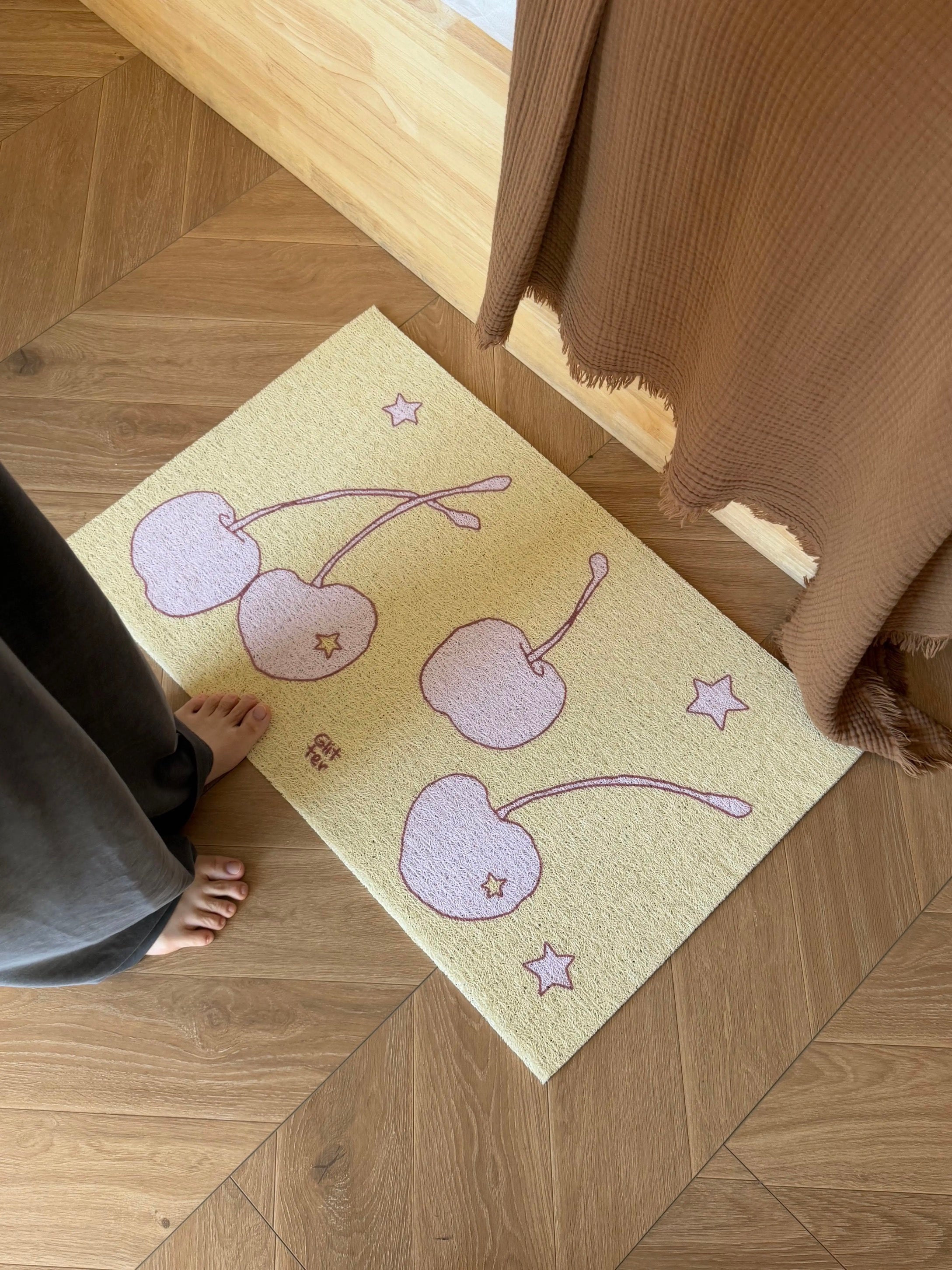 A rectangular cream-colored textured rug with a pattern of three pink cherries and small pink stars, shown on a wooden herringbone floor next to a person's bare feet and a brown curtain.