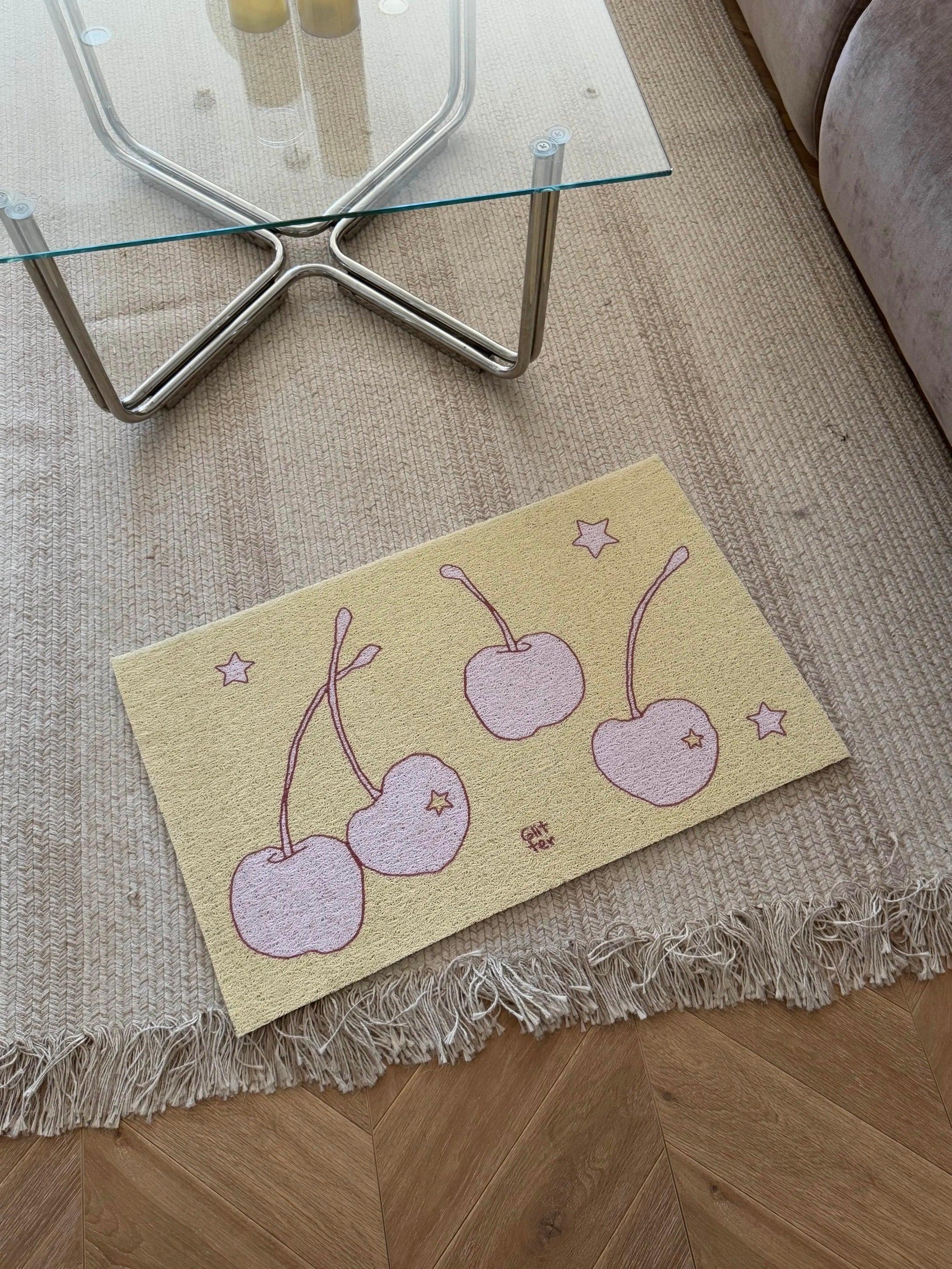 A rectangular beige rug with a fringed edge lies on a wooden floor in a living room setting. The rug features a whimsical pattern of three pink cherries with stems and small pink stars.