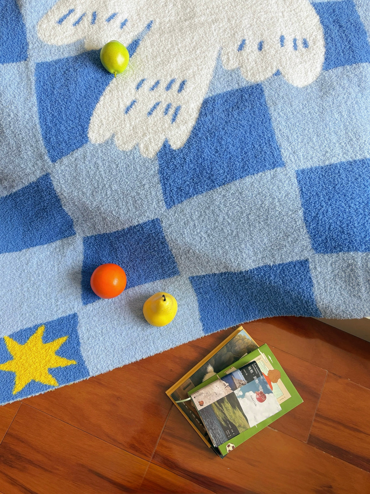 A blue and white checkered plush rug with a white dove motif and a yellow star, styled on a wooden floor with small toy fruits and a book.